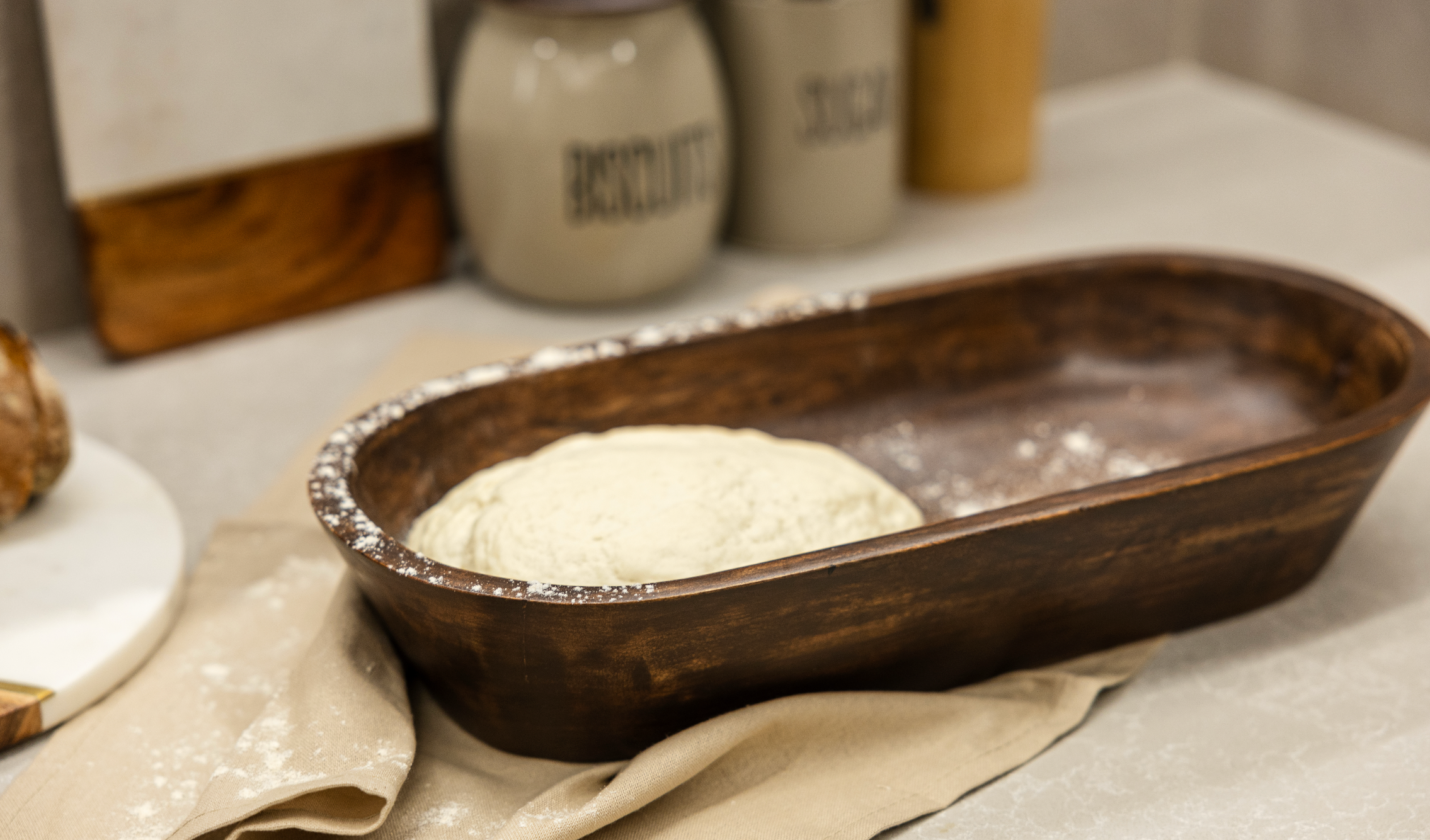 Wooden dough bowl with bread dough resting inside on a kitchen counter.