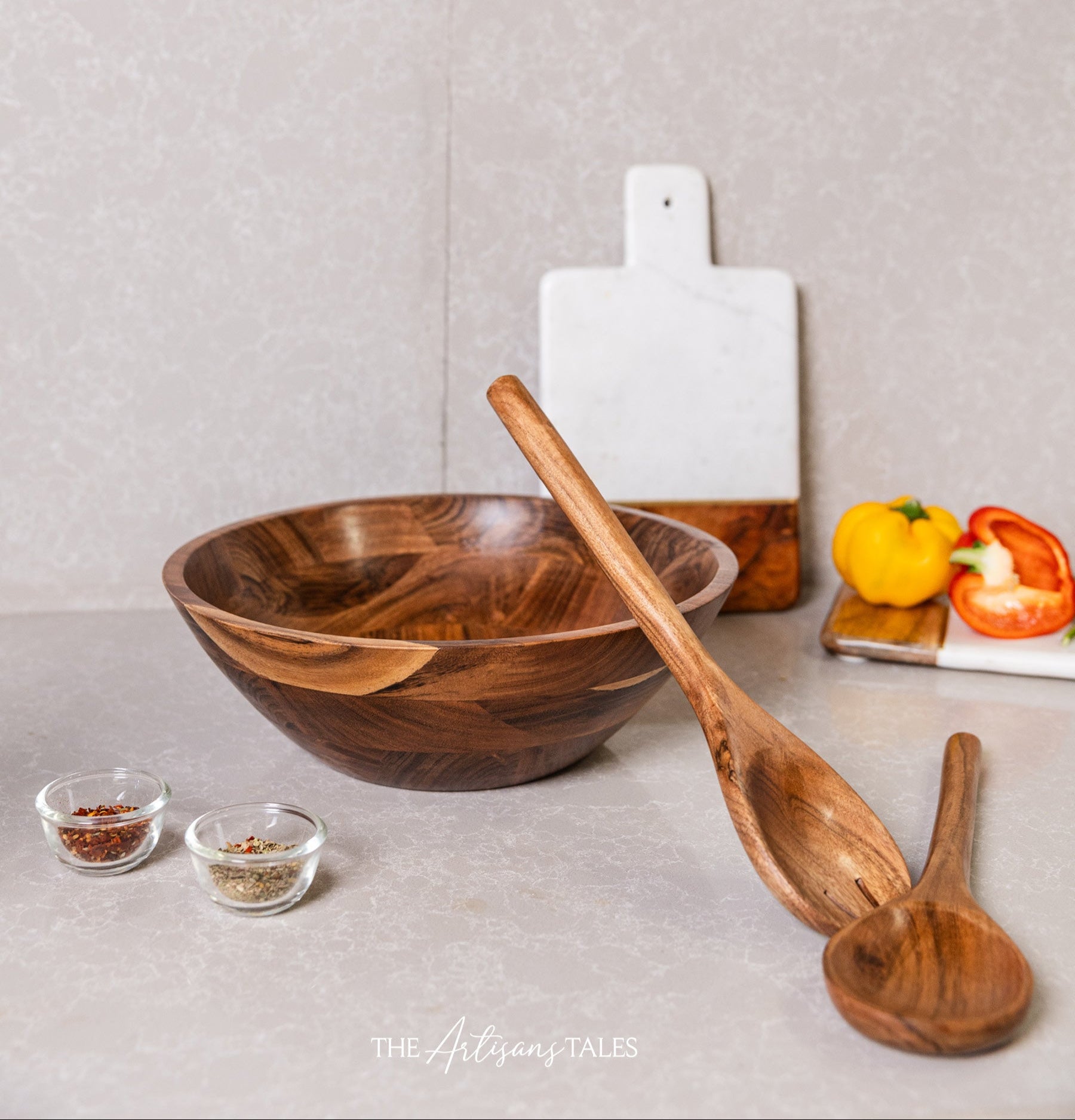 Wooden salad bowl with matching wooden serving spoons displayed on a kitchen counter with spices and vegetables.